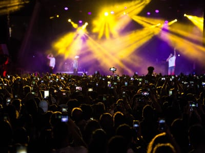 Concert crowd raising phones toward two performers on stage lit by yellow and purple spotlights