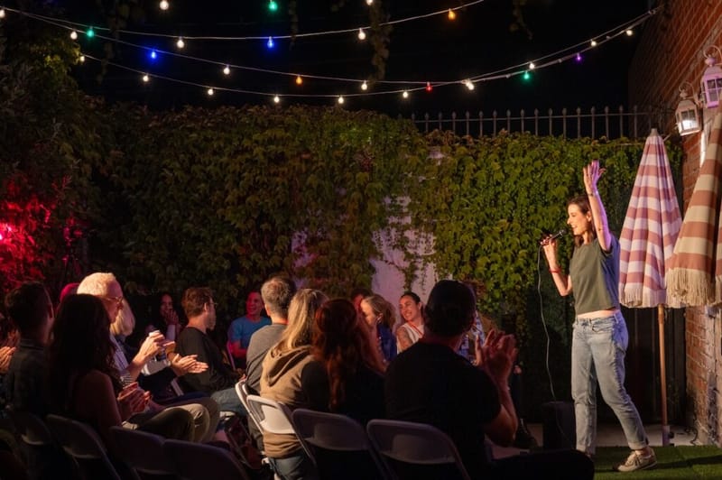 A comedian performs stand-up to a seated backyard audience under string lights at the Laughs on Larchmont comedy crawl.