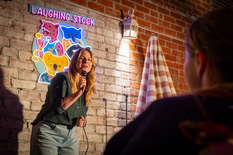 A comedian performs in front of a neon 'Laughing Stock' sign at the Laughs on Larchmont show.