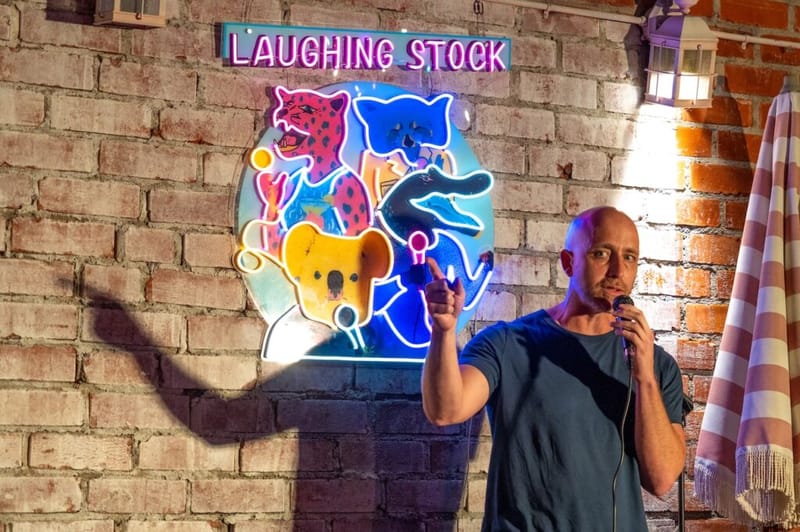 A comedian performs stand-up beside the neon 'Laughing Stock' sign at the Laughs on Larchmont show.