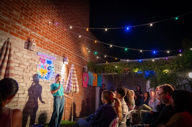 A comedian performs to a backyard crowd under string lights, with event t-shirts and a neon sign on the brick wall.