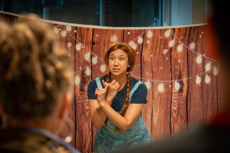 A character comedian in a red wig and overalls performs a sketch in front of a string-light backdrop.