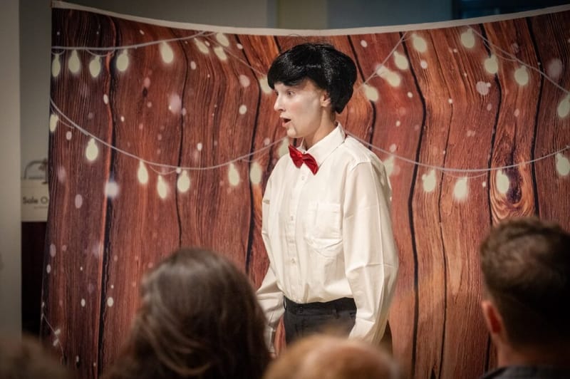 A performer in whiteface, a black wig, and a red bow tie does a character bit at the comedy crawl.