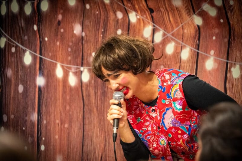 A character comedian with rosy clown cheeks performs energetically into the microphone.