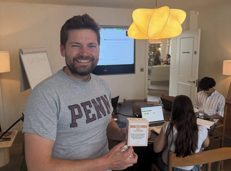 A tutor in a Penn t-shirt holds a box of SAT and ACT math flashcards at the London boot camp, students studying behind him.